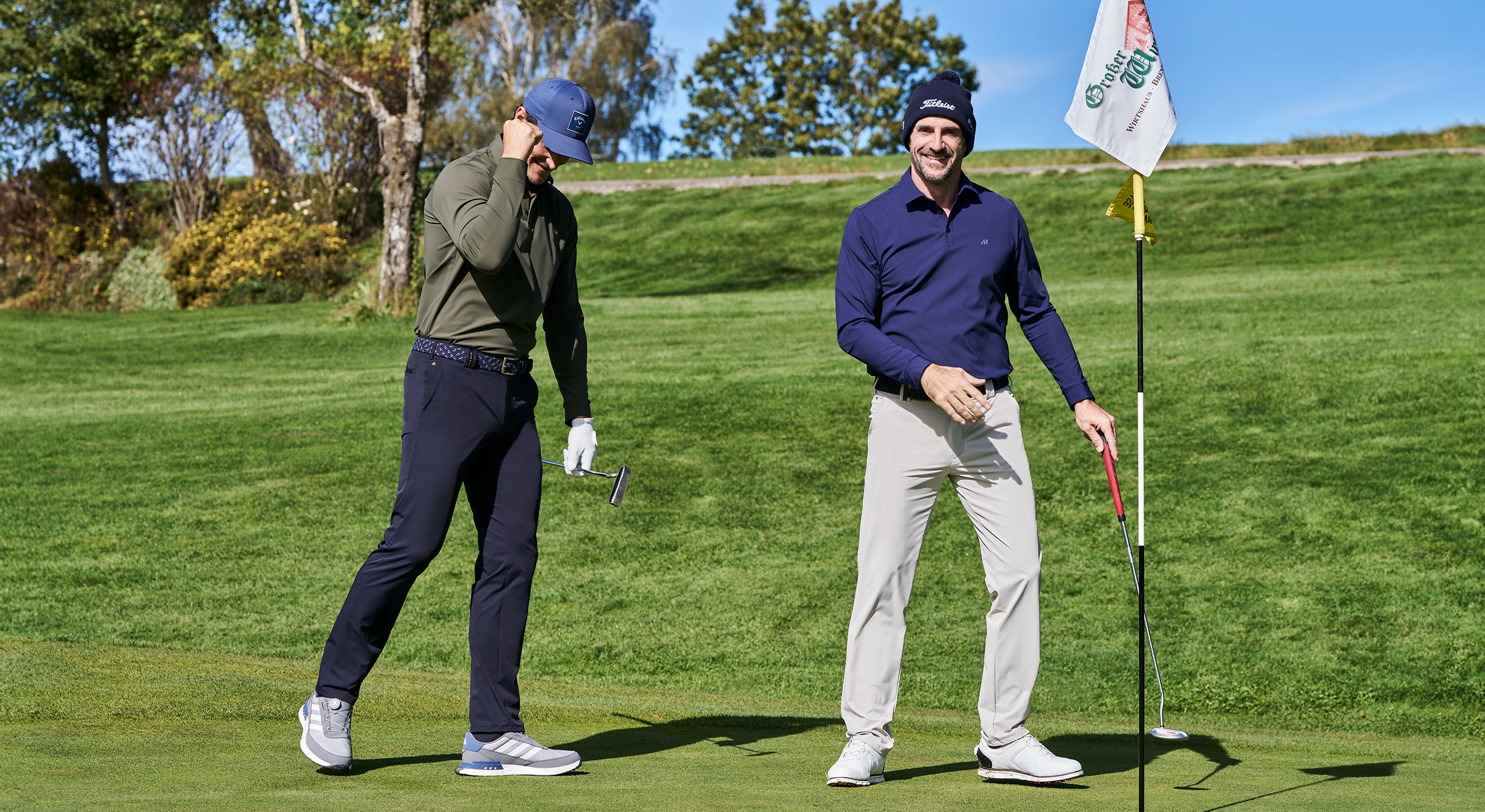 Two men on a golf course with one holding a golf club and the other standing next to a flag.